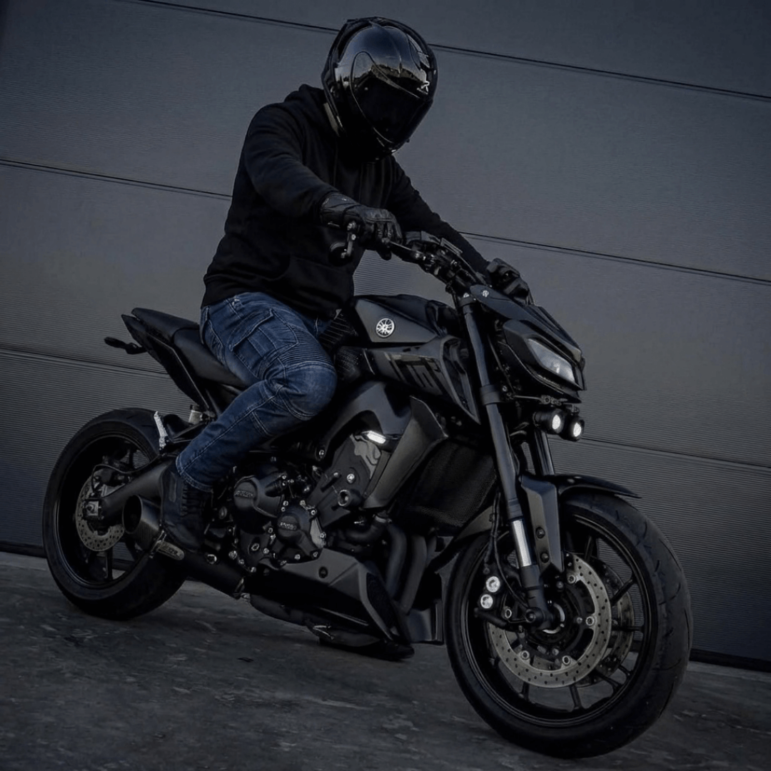 Rider in a black helmet and hoodie on a matte black motorcycle, wearing dark blue NORDRYK Redback motorcycle jeans, side-on shot against a metal wall