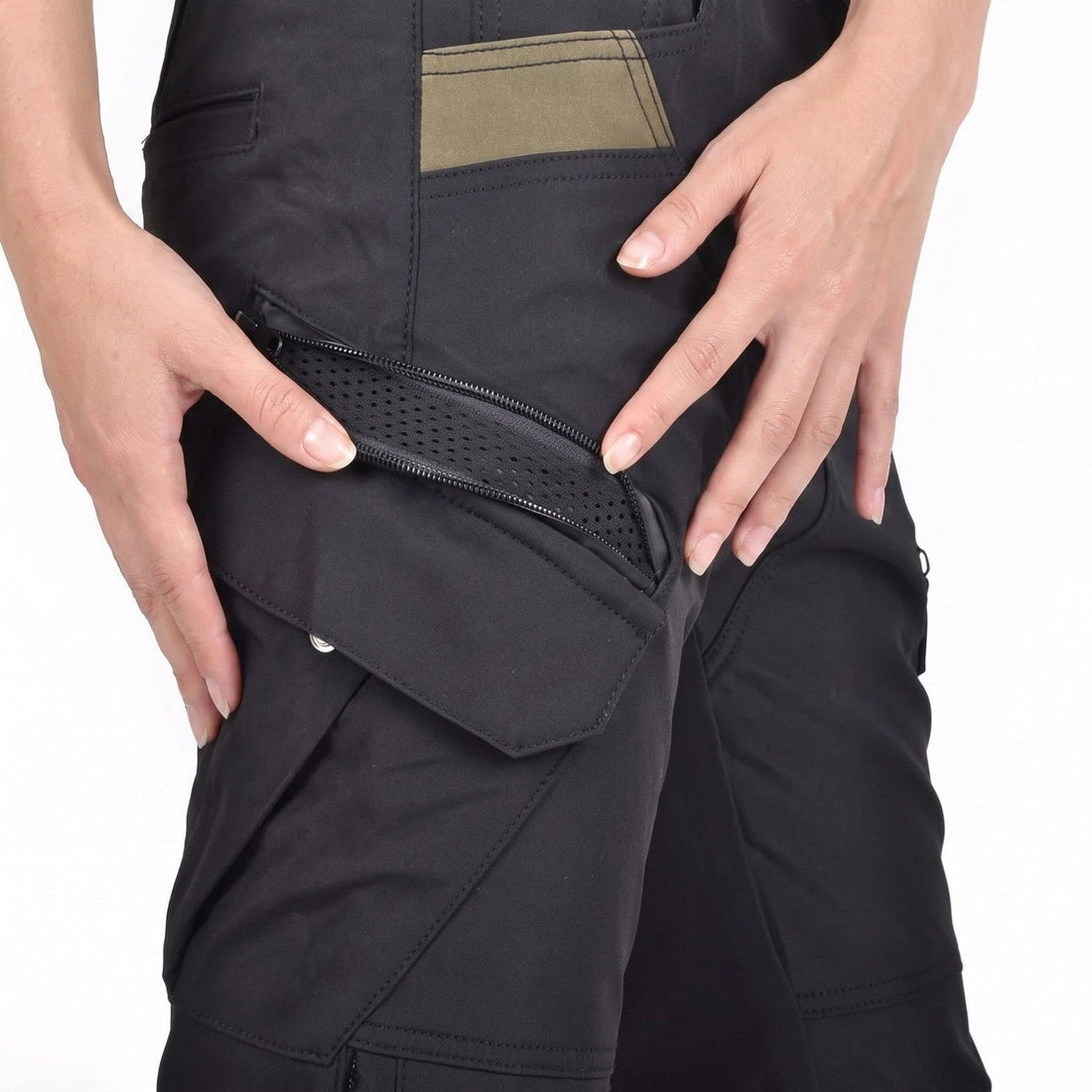 Two hands holding open a diagonal zip on the thigh pocket of black NORDRYK RoadVault cargo trousers, showing black perforated mesh underneath, with a flap pocket below and an olive panel above