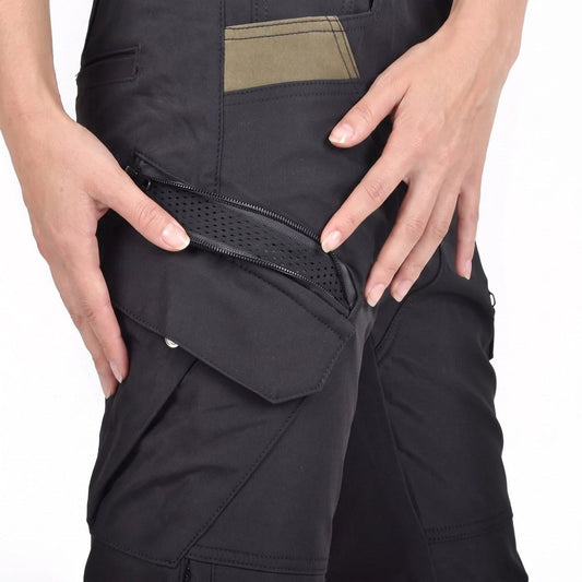 Two hands holding open a diagonal zip on the thigh pocket of black NORDRYK RoadVault cargo trousers, showing black perforated mesh underneath, with a flap pocket below and an olive panel above