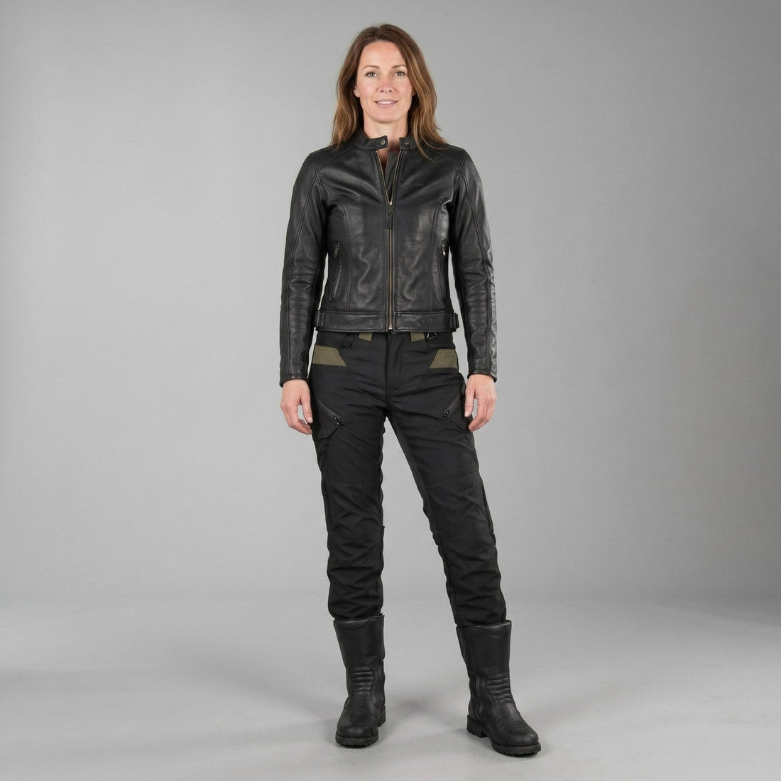 Woman in black NORDRYK RoadVault cargo trousers, a black jacket and tall black boots, standing against a plain grey studio wall