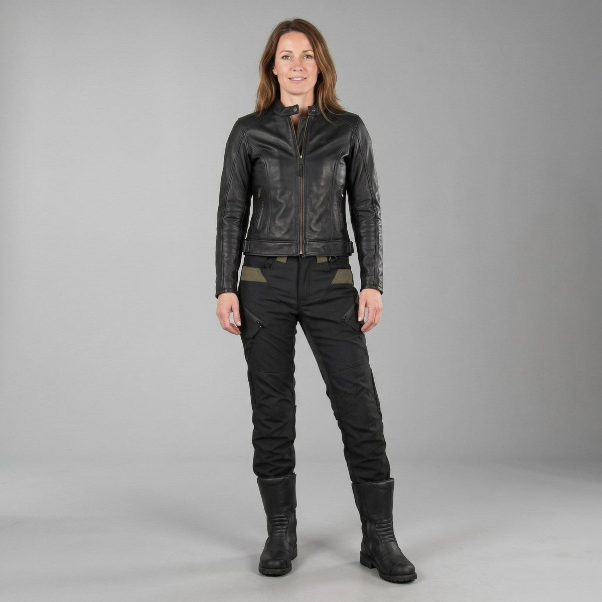 Woman in black NORDRYK RoadVault cargo trousers, a black jacket and tall black boots, standing against a plain grey studio wall